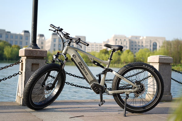 HJM Apex silver-gray full-suspension electric bike parked beside a waterfront